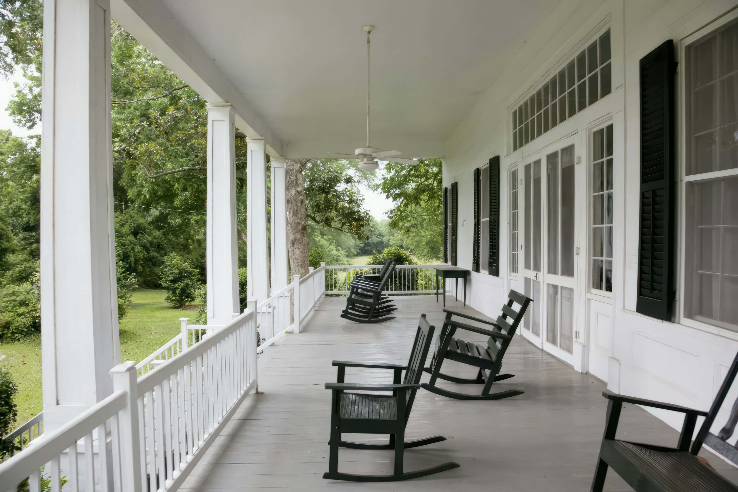 A long white porch lined with black rocking chairs, looking out over a green lawn and trees